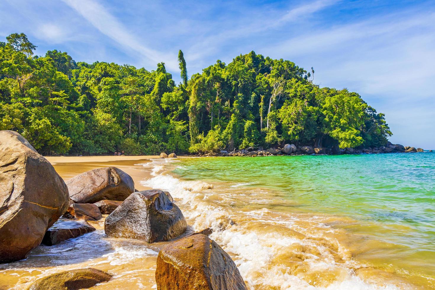ภาพวิวชายหาดที่มีหาดทรายสีทอง น้ำทะเลสีเขียวใส สีฟ้าของท้องฟ้า และโขดหินใหญ่สีเข้มในซีกซ้ายของภาพด้านหน้า มีป่าไม้เขียวชอุ่มหนาแน่นอยู่ริมชายหาด แสงแดดส่องสว่าง ทำให้น้ำทะเลและต้นไม้ดูสดใส สะท้อนบรรยากาศที่สงบและสวยงามตามธรรมชาติ เหมาะกับการพักผ่อนและเที่ยวชมธรรมชาติริมทะเล