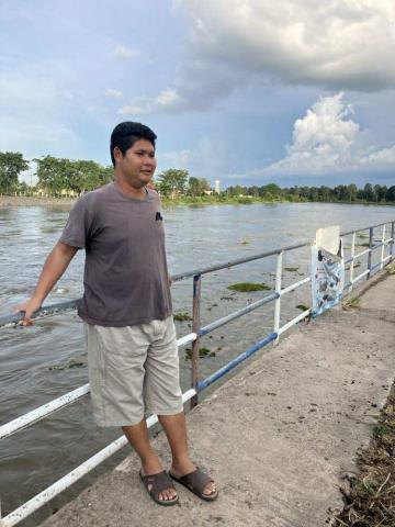 ผู้ชายคนหนึ่งยืนอยู่บนทางเดินริมแม่น้ำ เขาสวมเสื้อยืดสีเทา กางเกงขาสั้นสีครีม และรองเท้าแตะสีน้ำตาล มือขวาจับราวเหล็กที่เป็นรั้วกั้นริมแม่น้ำ ด้านหลังเป็นแม่น้ำกว้างและมีต้นไม้เขียวขจีอยู่ฝั่งตรงข้าม ท้องฟ้ามีเมฆมาก บรรยากาศดูผ่อนคลายและเป็นธรรมชาติ