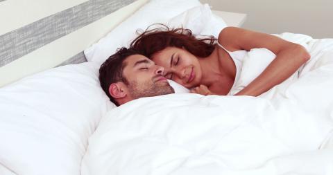 คู่รักนอนอยู่บนเตียง ทั้งสองคนกำลังหลับอยู่ใต้ผ้าห่มสีขาว ผู้หญิงนอนกอดผู้ชายจากด้านข้าง ดูมีความสุขและผ่อนคลาย ห้องดูสว่างและสะอาด เน้นความรู้สึกอบอุ่นและสบาย