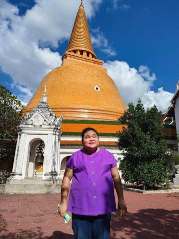 ในภาพนี้มีผู้หญิงคนหนึ่งยืนอยู่ด้านหน้าของเจดีย์ขนาดใหญ่ที่มีสีส้มทอง ซึ่งเป็นเจดีย์ที่มีลักษณะเป็นทรงระฆังและมีปลายแหลมสูงขึ้นไปบนท้องฟ้า ท้องฟ้าในภาพมีสีฟ้าสดใสและมีเมฆขาวลอยอยู่ ผู้หญิงคนนี้สวมเสื้อสีม่วงและกางเกงยีนส์ เธอกำลังยิ้ม ด้านข้างของเจดีย์มีต้นไม้สีเขียวและอาคารสีขาวที่มีหลังคาสีส้มและเขียว