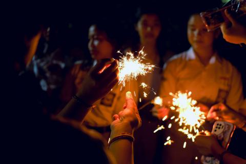 กลุ่มคนกำลังเล่นพลุเย็นในคืนเฉลิมฉลองปีใหม่ มีแสงไฟสีส้มจากพลุเย็นส่องสว่างบนมือ หลายคนดูเหมือนจะเป็นวัยรุ่นหรือเด็กนักเรียน สวมชุดนักเรียนสีอ่อน ในมือบางคนถือของมีลายการ์ตูนหรือโทรศัพท์มือถือ ทุกคนดูมีความสุขและเพลิดเพลินกับการเล่นพลุ พลุเย็นกำลังส่องประกายแสงเป็นประกายเล็ก ๆ รอบ ๆ ตัวอยู่ในบรรยากาศรื่นเริง