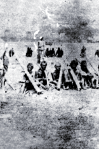 ภาพขาวดำกลุ่มผู้นำและชาวบ้านที่ถูกจับกุม ในฐานะกบฏผู้มีบุญ ณ ทุ่งศรีเมือง จังหวัดอุบลราชธานี เมื่อปี พ.ศ. 2444 ผู้ถูกจับกุมหลายคนถูกใส่ขื่อคา และนั่งอยู่บนพื้นดิน มีทหารสยามยืนควบคุมอยู่ด้านหลัง