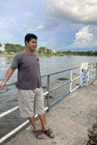 ผู้ชายคนหนึ่งยืนอยู่บนทางเดินริมแม่น้ำ เขาสวมเสื้อยืดสีเทา กางเกงขาสั้นสีครีม และรองเท้าแตะสีน้ำตาล มือขวาจับราวเหล็กที่เป็นรั้วกั้นริมแม่น้ำ ด้านหลังเป็นแม่น้ำกว้างและมีต้นไม้เขียวขจีอยู่ฝั่งตรงข้าม ท้องฟ้ามีเมฆมาก บรรยากาศดูผ่อนคลายและเป็นธรรมชาติ