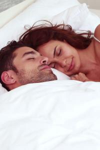 คู่รักนอนอยู่บนเตียง ทั้งสองคนกำลังหลับอยู่ใต้ผ้าห่มสีขาว ผู้หญิงนอนกอดผู้ชายจากด้านข้าง ดูมีความสุขและผ่อนคลาย ห้องดูสว่างและสะอาด เน้นความรู้สึกอบอุ่นและสบาย