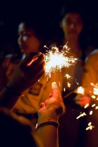 กลุ่มคนกำลังเล่นพลุเย็นในคืนเฉลิมฉลองปีใหม่ มีแสงไฟสีส้มจากพลุเย็นส่องสว่างบนมือ หลายคนดูเหมือนจะเป็นวัยรุ่นหรือเด็กนักเรียน สวมชุดนักเรียนสีอ่อน ในมือบางคนถือของมีลายการ์ตูนหรือโทรศัพท์มือถือ ทุกคนดูมีความสุขและเพลิดเพลินกับการเล่นพลุ พลุเย็นกำลังส่องประกายแสงเป็นประกายเล็ก ๆ รอบ ๆ ตัวอยู่ในบรรยากาศรื่นเริง