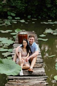 คู่รักนั่งอยู่บนสะพานไม้พาดข้ามสระน้ำที่มีใบบัวจำนวนมาก สองคนนั่งใกล้ชิดกันในท่าทางที่ดูอบอุ่น ฝ่ายหญิงใส่ชุดกระโปรงสีอ่อนและรองเท้าผ้าใบสีขาว ฝ่ายชายสวมเสื้อเชิ้ตแขนสั้นและกางเกงขาสั้น สภาพแวดล้อมรอบๆ เป็นธรรมชาติ เขียวขจี ดูสงบและผ่อนคลาย สะพานเป็นไม้ซุงเล็กๆ วางเรียงกันอย่างเรียบง่าย บรรยากาศโดยรวมดูผ่อนคลายและโรแมนติกมาก