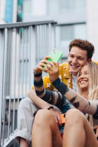 ชายหญิงคู่หนึ่งนั่งอยู่บนบันไดข้างนอกอาคาร พวกเขายิ้มและกำลังถ่ายเซลฟี่ด้วยโทรศัพท์มือถือที่มีเคสสีเขียว ฝั่งผู้ชายสวมเสื้อเชิ้ตแขนสั้นสีเหลืองลายดอกไม้ ส่วนผู้หญิงไว้ผมยาว สวมเสื้อแขนยาวลวดลายตัวสีเบจและน้ำเงิน ทั้งสองดูมีความสุขและผ่อนคลาย ข้างหลังเป็นราวเหล็กและอาคารสูงสมัยใหม่