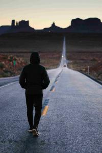 ภาพมุมกว้างของถนนเส้นยาวที่ทอดยาวไปไกลจนสุดสายตา ท่ามกลางหุบเขาและทะเลทราย มีคนหนึ่งคนกำลังเดินอยู่กลางถนน หันหลังให้กล้องและมุ่งหน้าไปยังดวงอาทิตย์ที่กำลังตกดินแสงสีทอง สื่อถึงความมุ่งมั่นที่ปนความสงบ