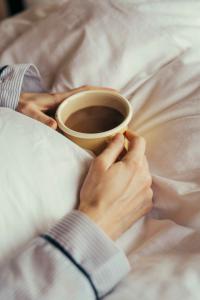 ภาพบุคคลนอนอยู่บนเตียง สวมชุดนอนที่ดูสบาย ในมือประคองแก้วกาแฟไว้ ใบหน้าดูผ่อนคลายแต่สื่อถึงพฤติกรรมการบริโภคเครื่องดื่มบนเตียง