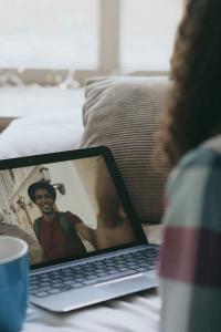 หญิงสาวนั่งอยู่บนเตียงอย่างผ่อนคลาย ขณะพูดคุยกับคนรักผ่านหน้าจอแล็ปท็อปที่วางอยู่ข้างแก้วกาแฟ เธอสวมเสื้อเชิ้ตลายสก็อตสีฟ้า-ชมพู ผมยาวและหยักศก ฝ่ายชายในหน้าจอสวมเสื้อยืดสีแดงและหมวกกำลังยิ้มและถ่ายวิดีโอจากถนนข้างอาคารสีขาว สะท้อนถึงความสัมพันธ์ทางไกลที่อบอุ่น