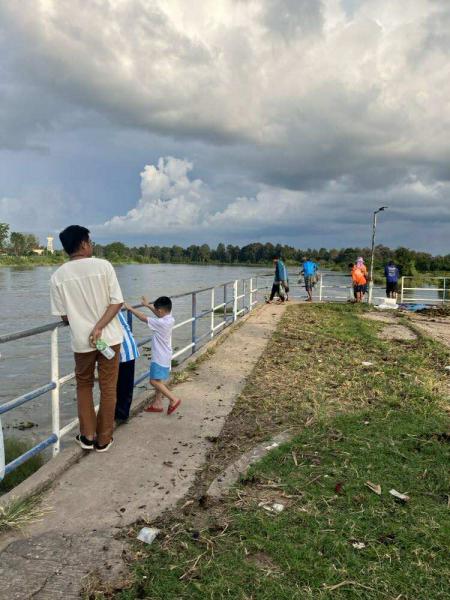 ทางเดินริมแม่น้ำ มีราวเหล็กกั้นข้างทางอยู่ทั้งสองฝั่ง คนกลุ่มหนึ่งกำลังยืนหรือเดินเล่นบนทางเดินนี้ ส่วนใหญ่ดูเหมือนมาชมวิว บางคนแต่งกายลำลอง เด็กชายคนหนึ่งกำลังยกแขนขึ้นเล่นหรือชี้อะไรบางอย่าง บริเวณข้างๆ เป็นสนามหญ้ามีเศษขยะและใบไม้อยู่ประปราย ฟ้าในภาพมีเมฆหนาทึบบางส่วนให้บรรยากาศเหมือนฝนใกล้ตก ฉากหลังมีต้นไม้และน้ำในแม่น้ำที่ค่อนข้างนิ่ง