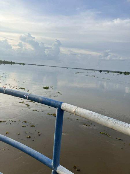 ภาพผืนน้ำกว้างใหญ่ ด้านหน้ามีราวเหล็กทาสีฟ้าขาว น้ำในภาพดูนิ่ง มีเศษวัชพืชหรือพืชน้ำลอยอยู่ประปราย ท้องฟ้ามีเมฆมาก สีโทนอ่อน ดูสงบและเป็นธรรมชาติ สะท้อนความรู้สึกเงียบสงบของบรรยากาศริมแม่น้ำหรือทะเลสาบ