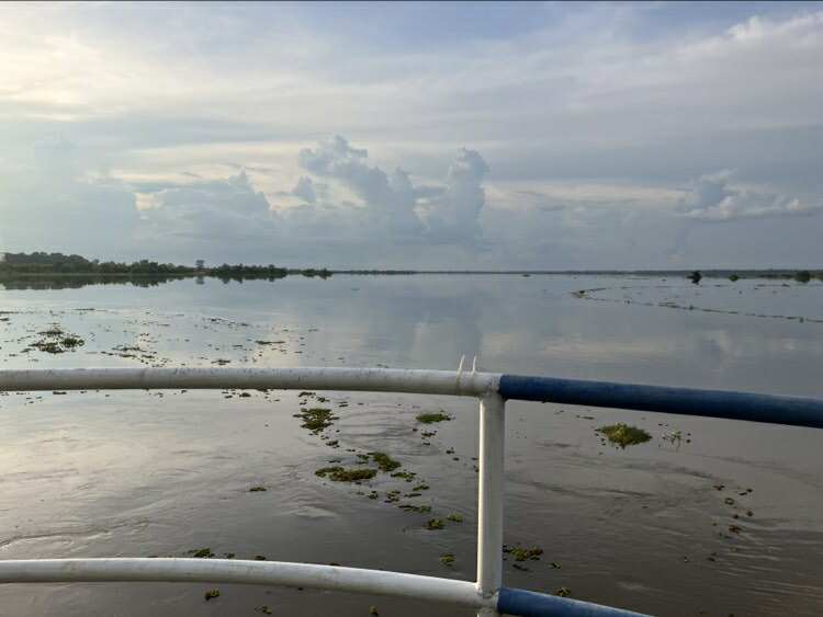 ภาพแม่น้ำกว้างใหญ่ในยามเย็น เห็นท้องฟ้าสีเทาอ่อนกับก้อนเมฆหนาแน่น มีราวเหล็กทาสีขาวกับน้ำเงินอยู่ด้านหน้า ขอบฟ้าด้านหลังเรียบโล่ง มีผักตบชวาลอยกระจายอยู่ในน้ำ บรรยากาศสงบเงียบและดูผ่อนคลาย