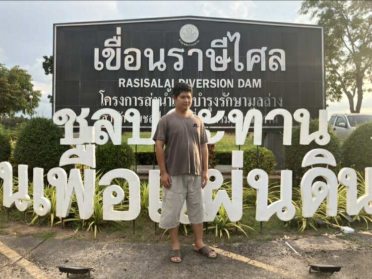 ภาพชายคนหนึ่งยืนอยู่หน้าป้ายขนาดใหญ่ ที่มีข้อความว่า “เขื่อนราษีไศล RASISALAI DIVERSION DAM โครงการส่งน้ำและบำรุงรักษามาตล่าง” อยู่ด้านหลัง ข้อความบนป้ายเด่นชัดและเป็นสีขาวบนพื้นสีดำ ส่วนชายคนนั้นสวมเสื้อยืดสีเทา กางเกงขาสั้นสีอ่อน และรองเท้าแตะ สภาพแวดล้อมโดยรอบเป็นธรรมชาติ มีต้นไม้และพุ่มไม้ด้านข้าง อากาศดูเหมือนช่วงเย็นหรือแดดร่ม